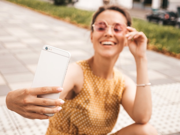 Closeup portrait of beautiful smiling brunette girl in summer hipster yellow dress. model taking selfie on smartphone.woman making photos in warm sunny day in the street in sunglasses