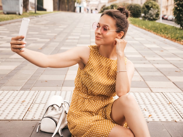 Closeup portrait of beautiful smiling brunette girl in summer hipster yellow dress. Model taking selfie on smartphone.Woman making photos in warm sunny day in the street in sunglasses
