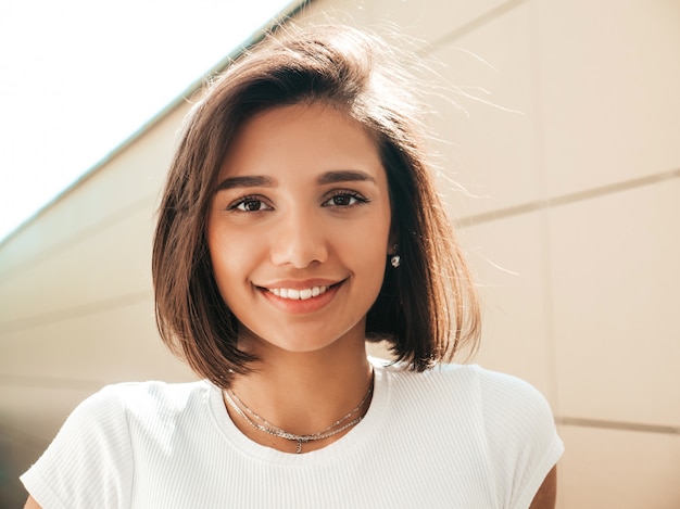 Il ritratto del primo piano di bello modello castana sorridente si è vestito in vestiti dei pantaloni a vita bassa dell'estate. ragazza d'avanguardia che posa nei precedenti della via. divertimento donna divertente e positiva