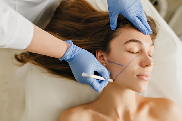 Free photo closeup portrait beautiful woman during cosmetology procedures, rejuvenation in beauty salon. dermatology procedure , painting eyebrows, hands in blue glows, at work, healthcare, botox