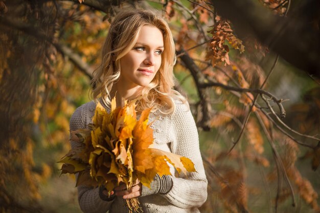 Closeup portrait of blond lady posing for photographer with a bouquet from maple leaves in the autumn forest