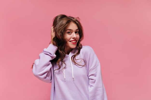 Closeup portrait of brunette curly woman touching her hair Pretty young girl in purple hoodie smiles and looks into camera on pink background