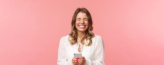 Closeup portrait of carefree tender lovely blond girl in white dress laughing over funny joke or message holding mobile phone close eyes and giggle at something hilarious pink background