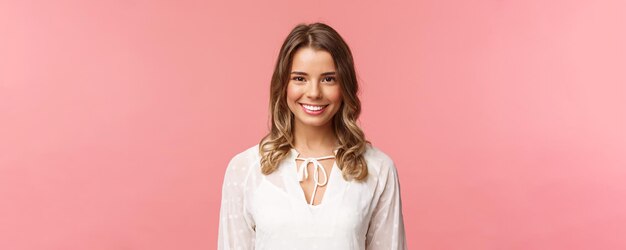 Closeup portrait of charming blond girl in lovely white dress with beaming smile looking at camera feeling happy and cheerful enjoying sunny romantic spring days pink background