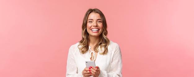 Closeup portrait of charming lovely blond girl using mobile phone texting friend order in online store using smartphone application laughing and smiling pleased stand pink background