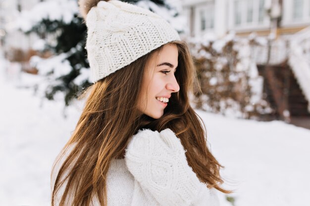 Очаровательная молодая женщина портрета крупного плана в белых шерстяных перчатках, связанной шляпе, длинных волосах брюнет, наслаждаясь погодой холодной зимы на улице. Улыбка в сторону, настоящие позитивные эмоции, веселое настроение.