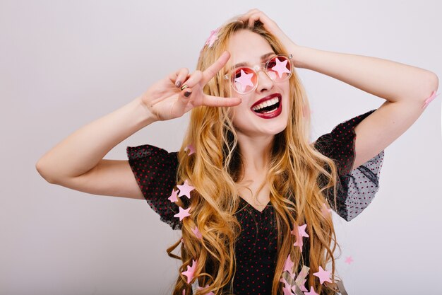 Closeup portrait of cheerful girl with blonde curly hair having great time at party, having fun, celebrating, showing peace. She wearing black dress, pink stylish glasses. Isolated..