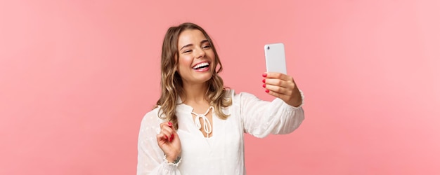 Closeup portrait of cheerful upbeat smiling blond girl wearing white dress laughing as record video calling friend on mobile application taking photo selfie on smartphone pink background