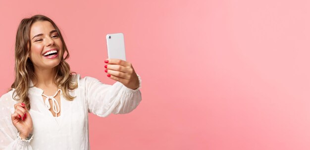 Closeup portrait of cheerful upbeat smiling blond girl wearing white dress laughing as record video calling friend on mobile application taking photo selfie on smartphone pink background
