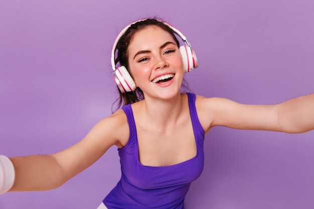 Closeup portrait of cheerful woman in fitness top and headphones taking selfie on purple wall