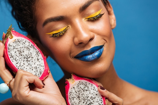 Il ritratto del primo piano della donna afro contenta con trucco luminoso che tiene la frutta di pitaya ha tagliato a metà prendendo piacere con gli occhi chiusi, sopra la parete blu