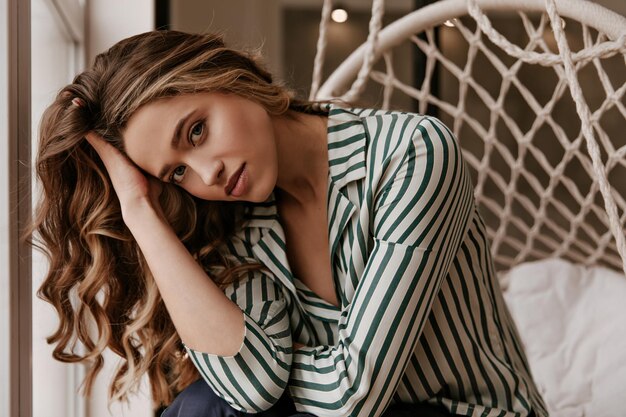 Closeup portrait of curly attractive lady in silk striped shirt looks into camera and sits in wick