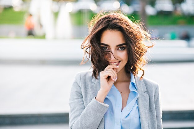 Closeup portrait of cute brunette girl with short hair walking in city.  She wears blue shirt, gray jacket.  She keeps hand on lips and smiling to camera.