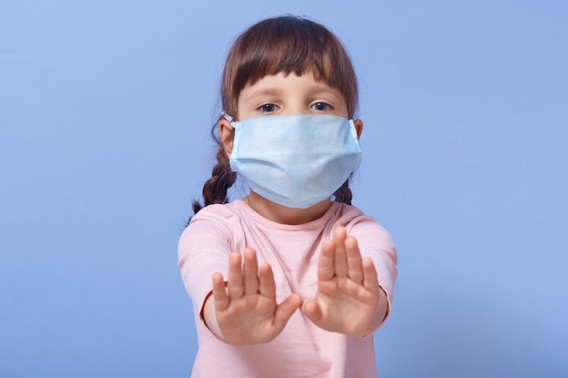 Closeup portrait of cute child wearing casual shirt and medical mask, female kid showing stop gesture with both palms