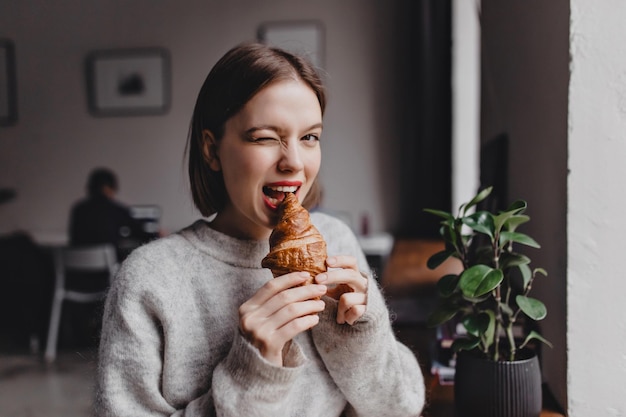 Ritratto del primo piano della ragazza bionda scura con il rossetto rosso che morde il croissant croccante contro lo spazio di coworking