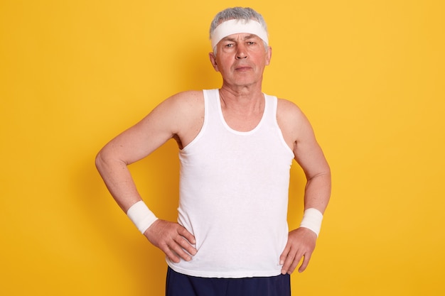 Closeup portrait of elderly man with hands on hips, wearing white sleeveless t shirt and headband doing sports