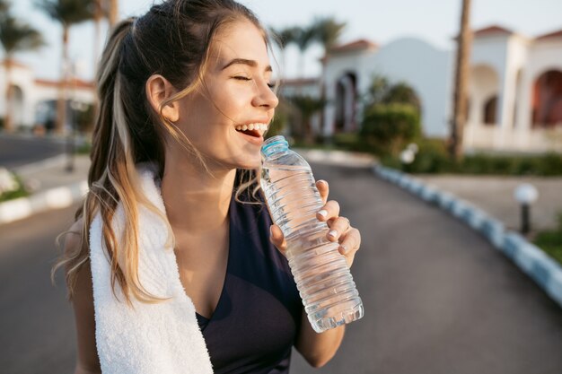 クローズアップの肖像画は、水のボトルと太陽に目を閉じて笑って幸せな若い女を興奮させた。魅力的なスポーツウーマン、夏を楽しみ、トレーニング、ワークアウト、幸福。