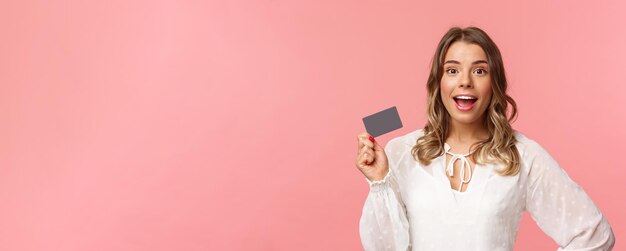 Closeup portrait of excited pretty young blond woman in white dress holding credit card showing peop