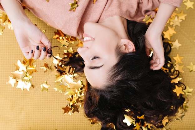 Free photo closeup portrait from above young joyful woman with cut curly hair having fun in golden tinsels on couch at home. lovely coisiness of pretty model, expressing positivity