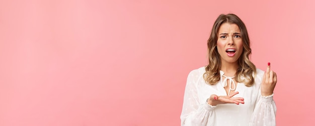 Closeup portrait of frustrated pissedoff complaining beautiful girl tired of waiting for proposal pointing at ring finger with bothered grimace standing pink background disappointed