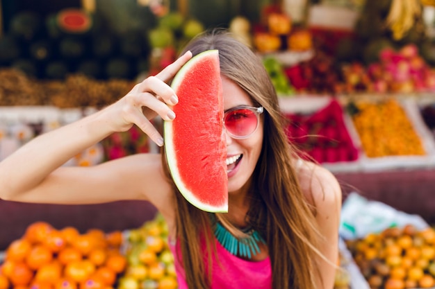 Closeup portrait of funny girl in pink sunglasses holding slice of watermelon on half face on tropical fruits market