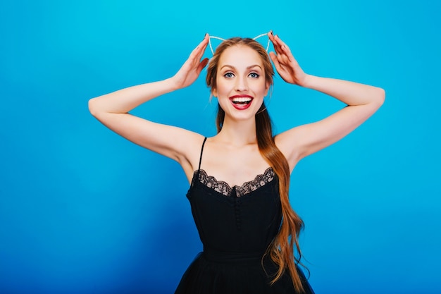 Free photo closeup portrait of gorgeous blonde ready for party, smiling and touching headband with cat ear in diamonds wearing beautiful black dress, bright makeup.