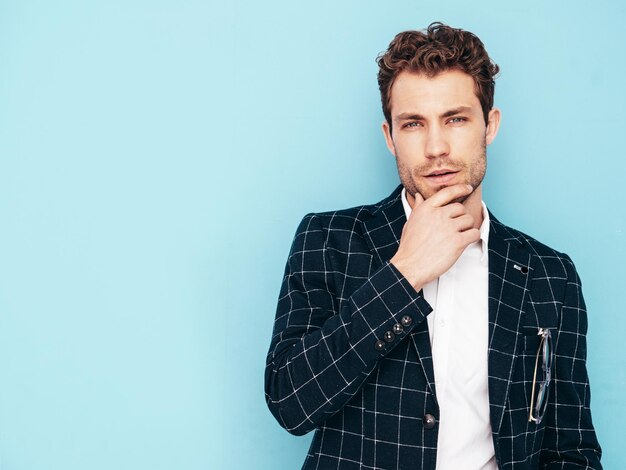 Closeup portrait of handsome confident stylish hipster lambersexual model Sexy modern man dressed in black elegant suit Fashion male posing in studio blue grey wall Touching his chin