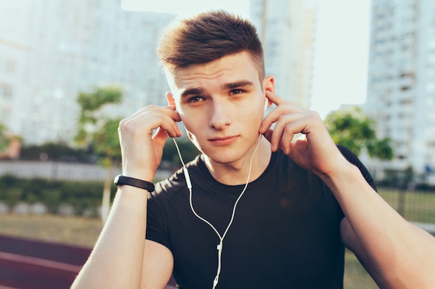Closeup portrait of handsome guy with a good body in the morning on stadium. He wears black T-shirt, put headphones to ears.