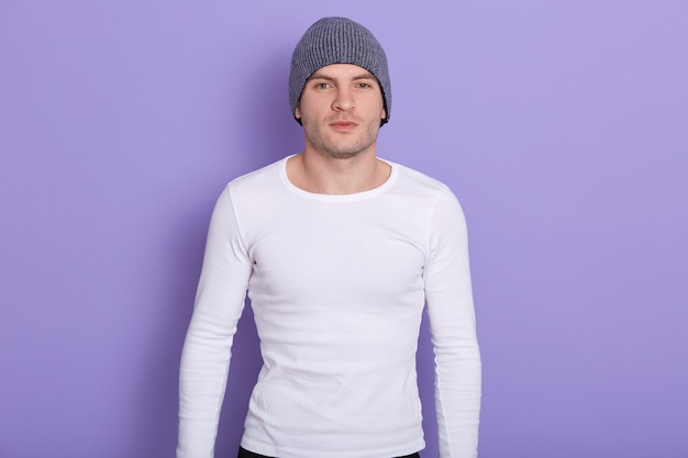 Free photo closeup portrait of handsome magnetic man standing isolated on lilac in, wearing grey hat and white sweatshirt, keeping strong fit. cold times concept.