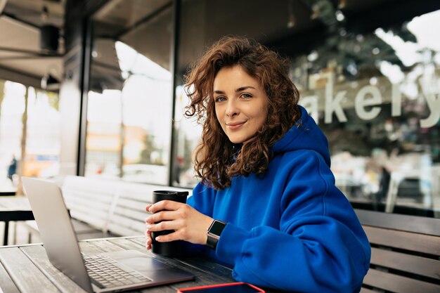 곱슬머리를 한 아름다운 여성의 클로즈업 초상화는 아침에 커피와 함께 노트북 작업을 하고 있습니다. 웃고 있는 수줍은 소녀의 야외 사진은 아침에 도시에서 원격으로 일하고 있습니다