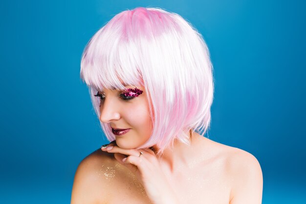 Closeup portrait joyful young woman with naked shoulders, pink haircut smiling to side . Brightful makeup with pink tinsels, sensitive, celebrating carnival, true emotions.