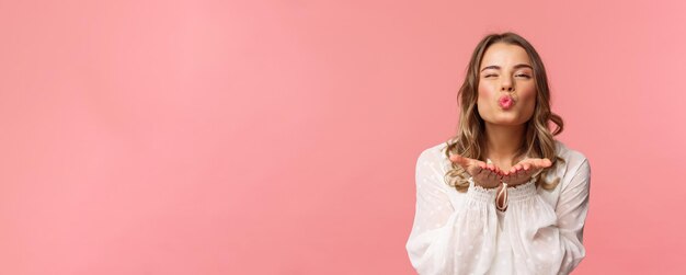 Closeup portrait of lovely feminine girlfriend enjoying spring fine day sending air kiss at camera winking and folding lips in mwah hold hands near mouth pink background