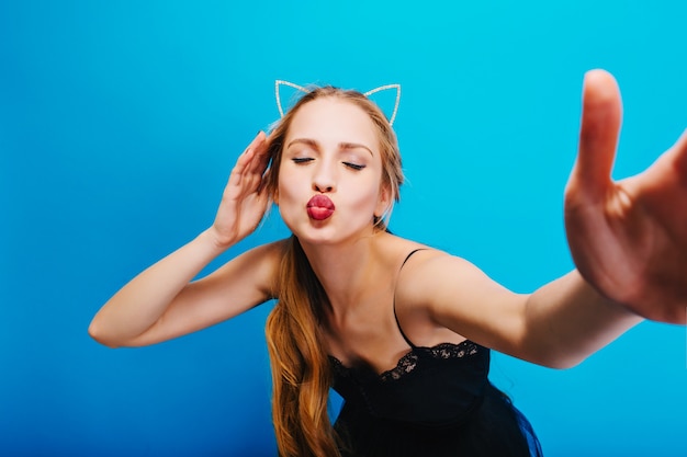 Foto gratuita ritratto del primo piano della ragazza adorabile che dà bacio, prendendo selfie, signora sulla festa. trucco luminoso con eyeliner. indossa un abito con pizzo, diadema di orecchie di gatto.