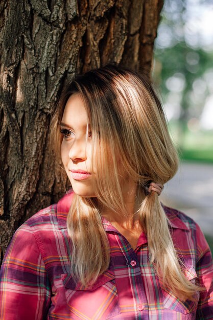 Closeup portrait photo of adult shining blonde woman with pigtails leaning on oak in park wearing ch...
