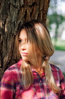 Closeup portrait photo of adult shining blonde woman with pigtails leaning on oak in park wearing ch...