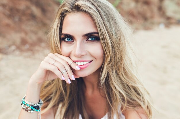 Closeup portrait of pretty blonde girl with long hair and blue eyes posing on rocky beach. She is smiling to the camera.