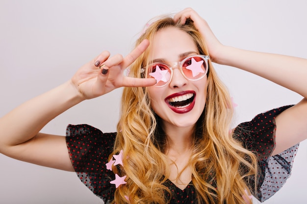 Closeup portrait of pretty blonde with curly hair enjoying time at party, celebrating, showing peace, smiling. She wearing black nice dress, pink glasses. 