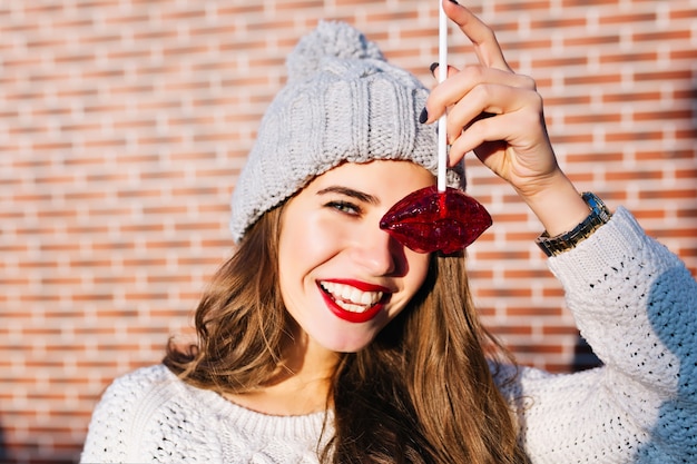 Closeup ritratto bella ragazza mora con capelli lunghi in cappello lavorato a maglia divertendosi con le labbra rosse caramello sulla parete esterna. lei sta sorridendo .