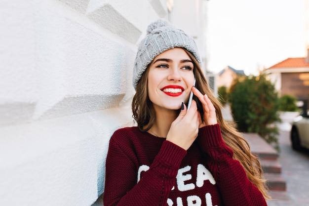 Closeup portrait pretty brunette with long hair in knitted hat and marsala sweater speaking on phone on street. She is smiling to side.