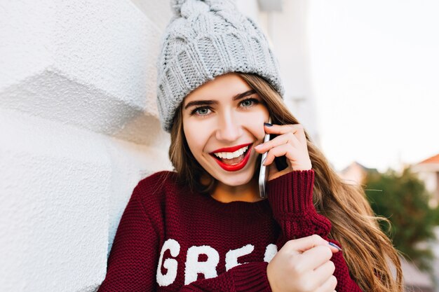 Closeup portrait pretty brunette with long hair in knitted hat and marsala sweater speaking on phone on street. She looks excited .