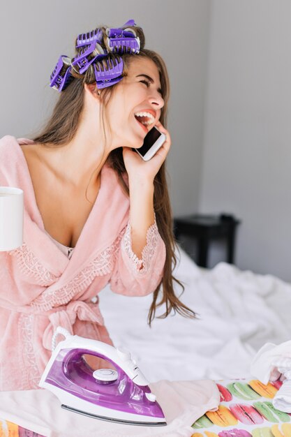 Closeup portrait pretty girl with long hair in pink bathrobe and curler on head at ironing clothes at home. She speaking on phone, laughing and looks enjoyed.