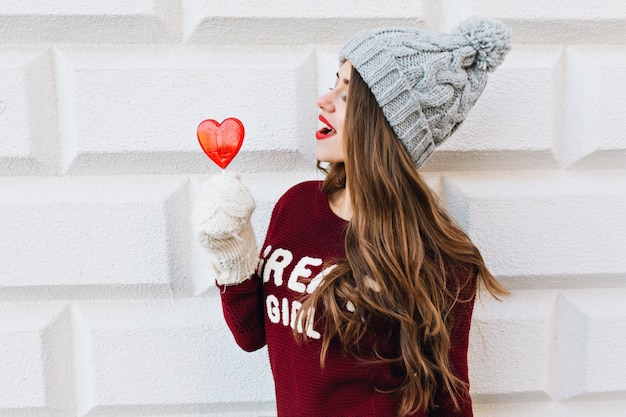 Портрет крупного плана милая маленькая девочка в свитере марсала и связанной шляпе на серой стене. У нее белые перчатки, она смотрит на леденец с красным сердцем в руке.