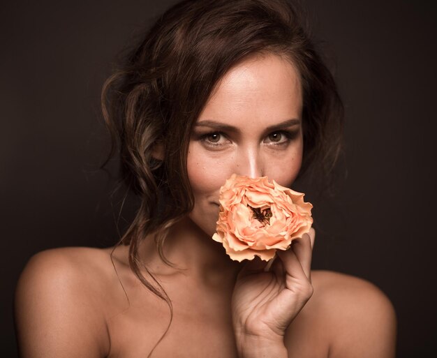 Крупным планом портрет профессиональной модели девушки, улыбающейся в темноте Красивая дама с каштановыми волосами смотрит в камеру и держит оранжевую розу возле рта