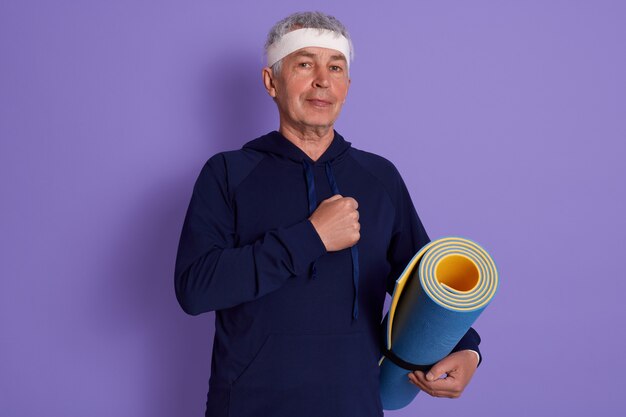 Closeup portrait of senior dresses blue hoody and white head band, keeping fist on his chest, holding yoga mat in hands, being photographed after physical exercise