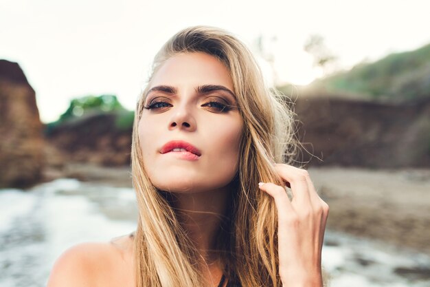 Closeup portrait of sexy blonde girl with long hair posing on rocky beach. She bites lips and looks to the camera.