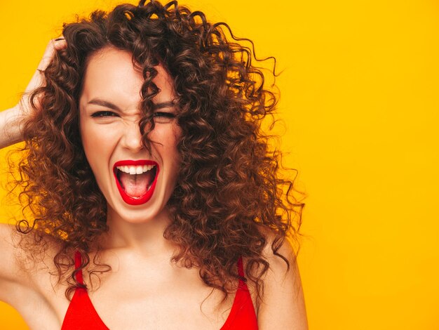 Closeup Portrait of sexy young beautiful smiling female Carefree woman isolated on yellow wall in studio Positive brunette model with afro curls hairstyle in sunglasses Screams and shouts