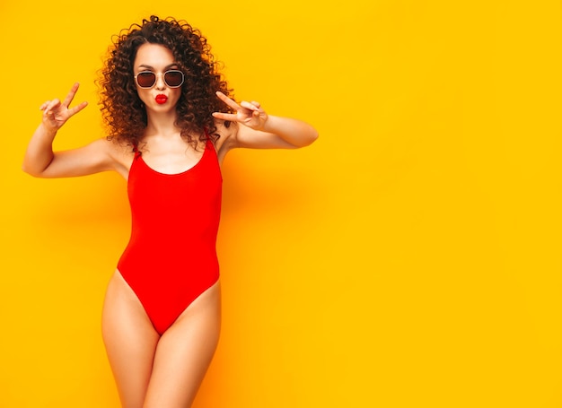 Closeup Portrait of sexy young beautiful smiling female Carefree woman isolated on yellow wall in studio Positive brunette model with afro curls hairstyle in sunglasses Shows peace sign