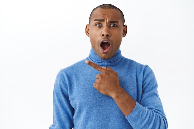 Closeup portrait of shocked and disappointed african american man complaining on something bad pointing finger left gasping open mouth and grimacing astounded of audacity white background