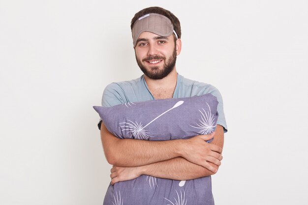 Closeup portrait of smiling bearded male embracing gray pillow, posing against white wall after waking up, wearing a sleep mask