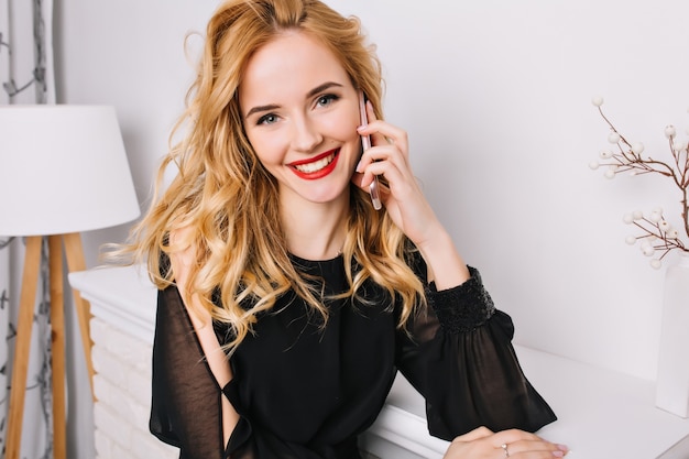 Closeup portrait of smiling blonde girl talking by phone, looking. Bright room with nice, modern white interior. Wearing stylish black dress. Floor lamp, fake fireplace.